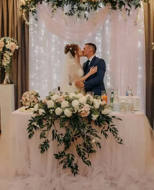 Novios en la mesa de honor con arreglo floral blanco y cortina de luces